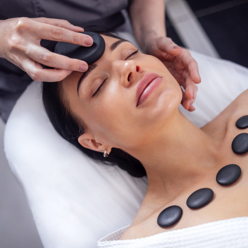 Close-up of a portrait of a young woman with her eyes closed receiving a facial massage with hot stones. A beautiful girl lies and relaxes during a spa procedure. Taking care of yourself concert.